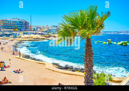 BUGIBBA, MALTA - JUNE 14, 2018: The stormy morning at Bugibba Perched Beach holds tourists on the coast, and the empty floating inflatable slides rock Stock Photo