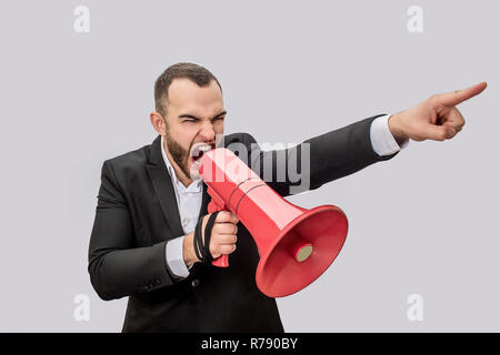Angry young man hold red megaphone in hand nad screma into it. He points forward. Guy is mad. Isolated on white background. Stock Photo