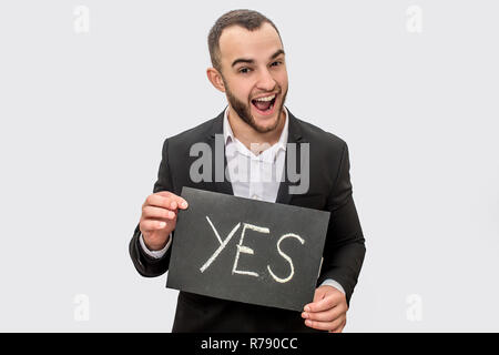 Happy young man in suit stands nd holds tablet with written word yes. Guy look straight on camera. He wears suit. Isolated on white background. Stock Photo