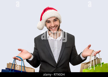 Cheerful and positive young man smiles on camera. He wears Christmas hat and black suit. Young man has shopping bags in both hands. Isolated on white background. Stock Photo