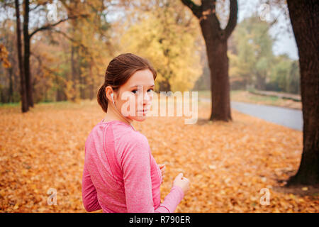 Young woman stands in autumn park and look back on camera. She is serious. Young woman has headphones in ears. She listening to music. Stock Photo