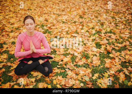 Calm and peaceful young woman sits in lotus position and meditating. She keeps eyes closed and ahnds together. Model is alone in park. Stock Photo
