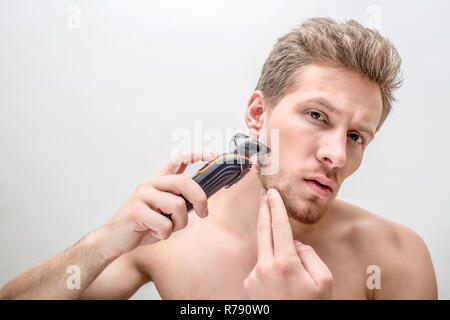 Serious young man shaving. He uses shaving machine. He works with cheek area. Isolated on white background. Stock Photo