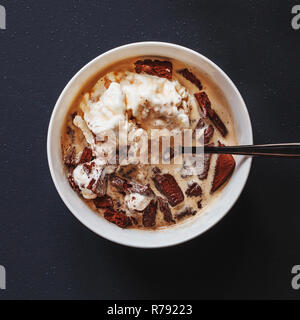 Plain white ceramic bowl filled with vanilla ice cream with liquer and crushed chocolate cookies placed on matte black painted table surface. Top view Stock Photo