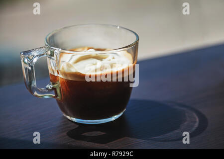Transparent glass cup of ice coffee with whipped cream placed on a dark wooden laminated table surface with blurredgray asphalt road behind a window g Stock Photo