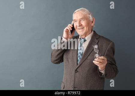 Senior man studio standing isolated on gray holding glasses talking on smartphone dreamful Stock Photo