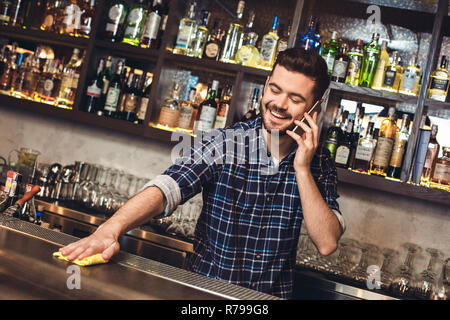 Young bartender standing at bar counter talking on smartphone cleaning table joyful Stock Photo