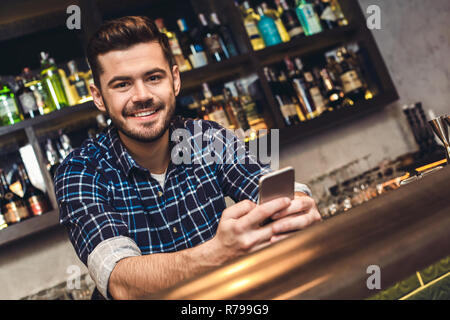 Young bartender leaning on bar counter holding smartphone happy Stock Photo