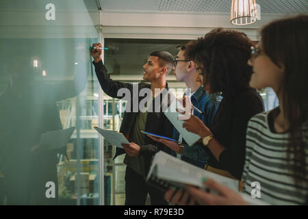 Male student writing an board in classroom with classmates standing around making notes. Student writing on board during class. Stock Photo