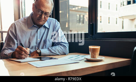 Senior businessman sitting at coffee shop table and writing notes on notepad. Man working from a modern cafe. Stock Photo