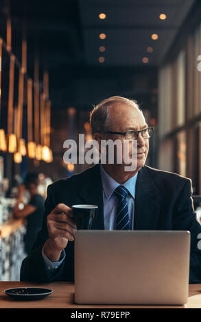 Vertical shot of senior businessman looking away and having coffee. Mature man in sitting at cafe table with laptop drinking coffee. Stock Photo