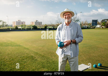 Smiling old man standing in a park with a boules in hand. Cheerful elderly man in hat standing in a play ground holding a boule. Stock Photo