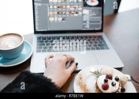 Top view of designers place. Womens hand using tochpad on laptop. Freelance working space. Notebook, cup of coffee and dessert on wooden table. Stock Photo