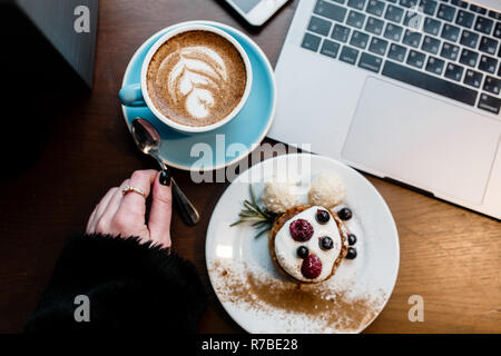 Top view of designers place. Womens hand keep spoon. Freelance working space. Notebook with mobile phone, cup of coffee and dessert on wooden table. Stock Photo