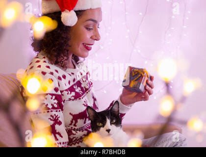 Happy Brazilian girl wearing christmas hat and seasonal sweater with adorable pet cat  holding gift box at home. festive decor with lights blurred. Stock Photo