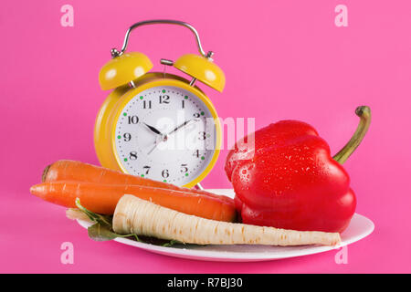 Vegetables on a plate and retro alarm clock isolated on pink background. Red pepper, carrots, parsnip and salad on plate. Chrono diet and nutrition Stock Photo