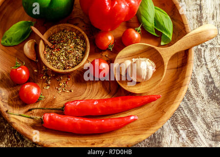 Fresh cherry tomatoes, garlic cloves, chili peppers, basil leaves, red and green capsicums, mix of dry spices and salt in a wooden pot. Stock Photo