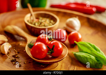 Fresh cherry tomatoes, garlic cloves, basil, mix of dry spices in wooden pot on wooden tray over wooden table, red and green capsicum. Stock Photo