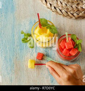 Cut cubes of red and yellow watermelon in glass jars with mint and canape picks over white and blue washed wooden table. Straw hat on the background Stock Photo