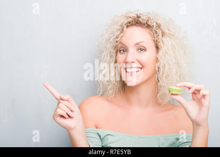 Young blonde woman over grunge grey background eating macaron very happy pointing with hand and finger to the side Stock Photo