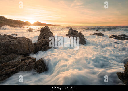 Long exposure of waves crashing onto rocks at sunset on the coast of the Balagne region of Corsica near Lozari with Ile Rousse in the distance
