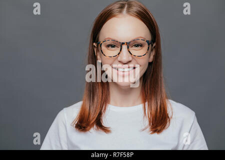 Headshot of pleasant looking dark haired young woman wears transparent glasses, dressed in casual clothes, has healthy skin, positive look, shows whit Stock Photo