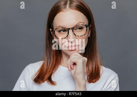 Thoughtful Caucasian woman with full lips, dark brown hair, looks thoughtfully aside, dressed in white clothes, being deep in thoughts, tries to make  Stock Photo