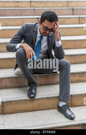 A sad looking young businessman sitting at the staircase looking down with stressed and depressed look gesture Stock Photo
