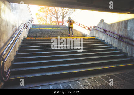 Tourist man in a hood with a large backpack climbs the stairs at dawn in an urban environment. Tourist in winter city concept Stock Photo