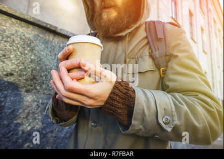 Paper cup of hot coffee in the hand of a bearded man in a jacket and a brown sweater. Warming drink in cold winter weather. Take away coffee concept.  Stock Photo