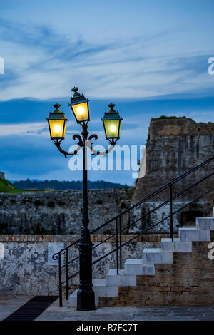 Vintage illuminated street lamp at dawn with three lights in front of ancient white stone steps of staircase found in the medieval fortress in Corfu I Stock Photo