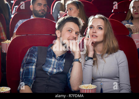 Cheerful couple having date in cinema. Smiling man talking it beautiful blonde girlfriend in gray. Cute pair watching interesting comedy and enjoying film together. Concept of free time of couple. Stock Photo