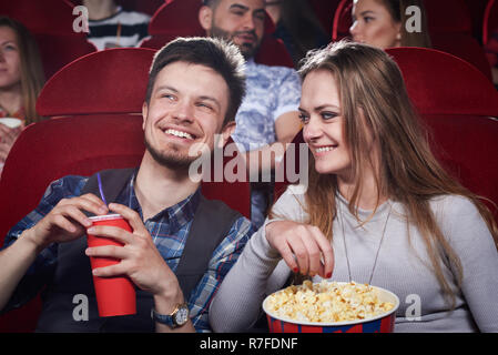 Happy cute couple eating popcorn and laughing at funny comedy in cinema theater. Attractive girl and handsome having romantic date and enjoying interesting movie. Concept of entertainment. Stock Photo