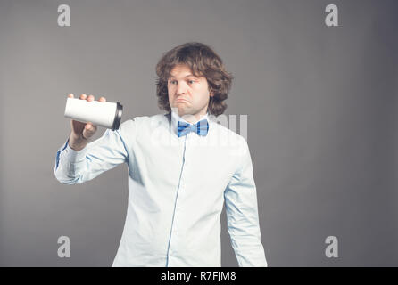 Upset guy with sad expression holding disposable paper cup of coffee upside down, looking disappointed, coffee is over. dressed in shirt and blue bow tie poses against grey background. Morning time. Stock Photo