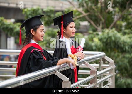 Two smiling female Asian students in graduation gown holding the diplomas with eyes looking forward Stock Photo