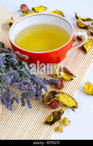Herbal tea in red cup, dry marjoram herbs and dry flower petals on bamboo mat background