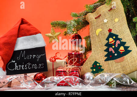 A blackboard with the text 'Merry Christmas' written in a chalk covered with a Santa Claus hat on a wooden board covered with snow next to the fir, gi Stock Photo