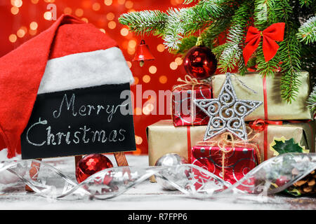 A blackboard with the text 'Merry Christmas' written in a chalk covered with a Santa Claus hat on a wooden board covered with snow next to the gifts. Stock Photo