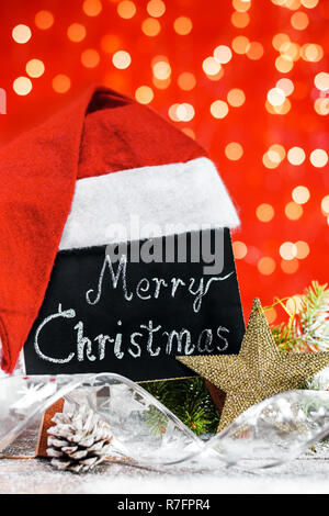 A table with the text 'Merry Christmas' written in a chalk covered with Santa's hat on a wooden board covered with snow with a branch of fir, pinecone Stock Photo