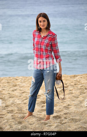 Beautiful russian brunette woman stands on the beach holding her photocamera, dressed in red chemise and blue jeans, the sea in the background Stock Photo