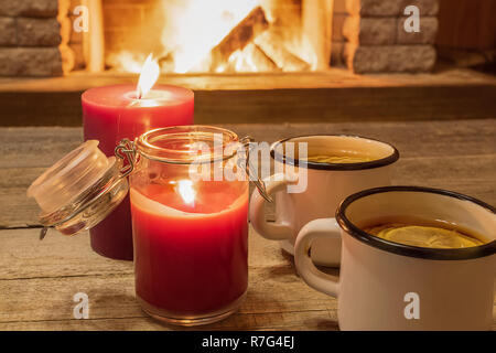 White enameled mugs with hot tea, and two candles , near cozy fireplace, in country house, winter vacation, horizontal. Stock Photo