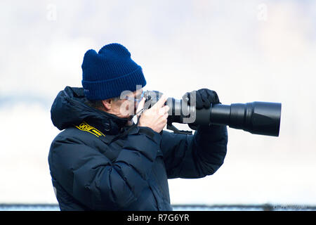 Photographer in winter pointing camera with large zoom lens with washed out background Stock Photo