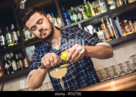 Young bartender standing at bar counter cutting lime zest into coktail concentrated Stock Photo