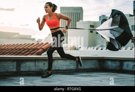 Female athlete doing drag running on rooftop. Side view of a fitness woman running on terrace of a building with a resistance parachute tied to her wa Stock Photo