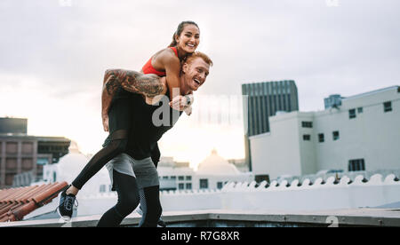 Athlete carrying a fitness woman on his back and walking on rooftop. Smiling fitness woman piggy riding on a man while training on rooftop. Stock Photo