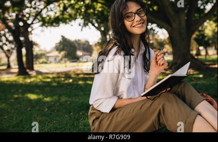 Beautiful young woman with glasses sitting at park writing in a book. Caucasian female writing in a book at park. Stock Photo