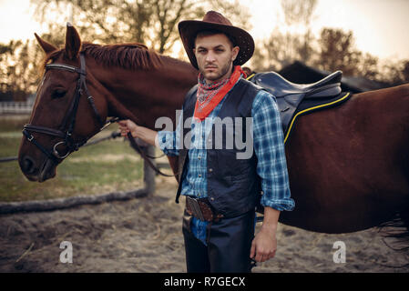 Brutal cowboy in jeans and leather jacket poses with horse on texas ranch, western. Vintage male person with animal, wild west Stock Photo