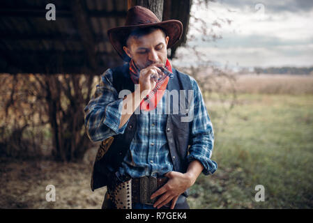 Brutal cowboy smokes a cigar, texas ranch on background, western. Vintage male person with gun on farm, wild west Stock Photo