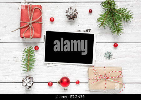 blank photo card in frame made of fir tree branches, decorations and gift boxes over white wooden background. christmas background. mock up. flat lay. Stock Photo