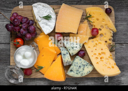 Assorted cheeses on wooden cutting board. Camembert cheese, cheddar, hard cheese slices, sof cheese, walnuts, grapes, tomatoes and rosemary. top view Stock Photo